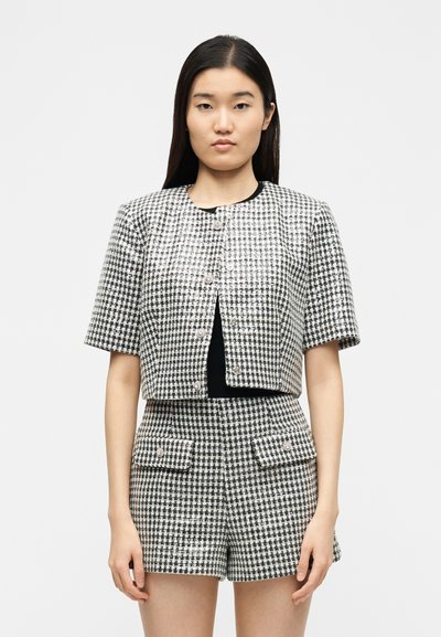 Femme portant une veste courte à manches courtes à carreaux noir et blanc et un short assorti taille haute, debout devant un fond uni.