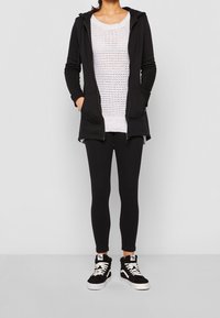 Persona che indossa una felpa nera con zip, un maglione bianco a maglia, leggings neri e scarpe alte bianco e nere, in piedi su uno sfondo semplice.