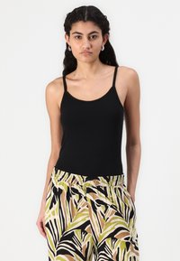Black tank top with thin straps, featuring a fitted design. Paired with patterned, loose-fitting trousers in green, brown, and black shades.
