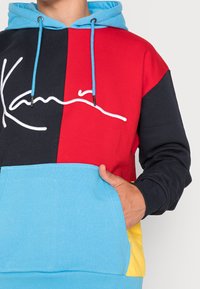 Sudadera con capucha en rojo, azul, negro y amarillo. Presenta un bolsillo frontal, capucha con cordón y un gráfico de firma en blanco. Tejido de mezcla de algodón.