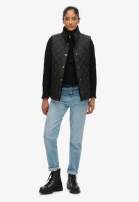 Czarna pikowana kamizelka z złotymi detalami, nałożona na czarny sweter. Jasnoniebieskie jeansy z podwiniętymi nogawkami i czarne sznurowane botki dopełniają stylizację.