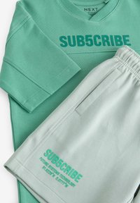 Green long-sleeve shirt and light green shorts with "SUB5CRIBE" printed on both garments and elastic waistband on shorts.