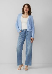 Lichtblauwe gebreide cardigan over een witte tanktop, gecombineerd met lichtblauwe wijde jeans en witte sandalen met hakken en banden.