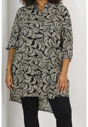Femme portant une chemise à manches longues boutonnée à motifs noir et blanc, un pantalon noir et des bracelets dorés à son poignet gauche.