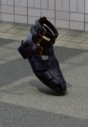 Donkerbruine leren sandaal met drie gespenriemen en een gevlochten voorkant, rechtop geplaatst op een bestrate ondergrond nabij een betegelde muur.