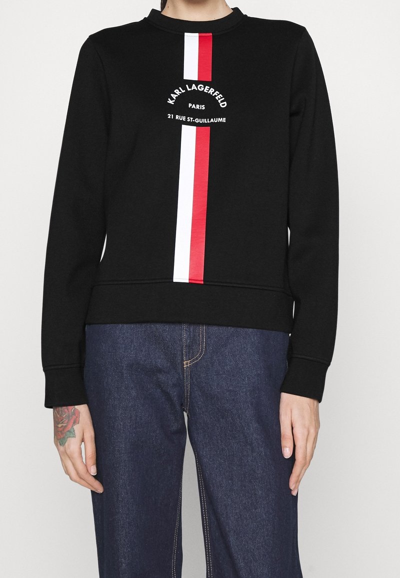 Personne portant un sweat-shirt noir avec des rayures verticales rouges et blanches et le texte "Karl Lagerfeld Paris 21 Rue St-Guillaume", associé à un jean bleu.