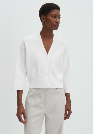 Vrouw die een witte cardigan met knopen en driekwart mouwen draagt, en een beige broek met hoge taille, staande tegen een effen lichte achtergrond.