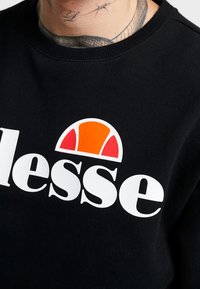 Primer plano de una sudadera negra con el logo de Ellesse en blanco y naranja, mostrando parte de un cuello tatuado sobre el cuello de la prenda.