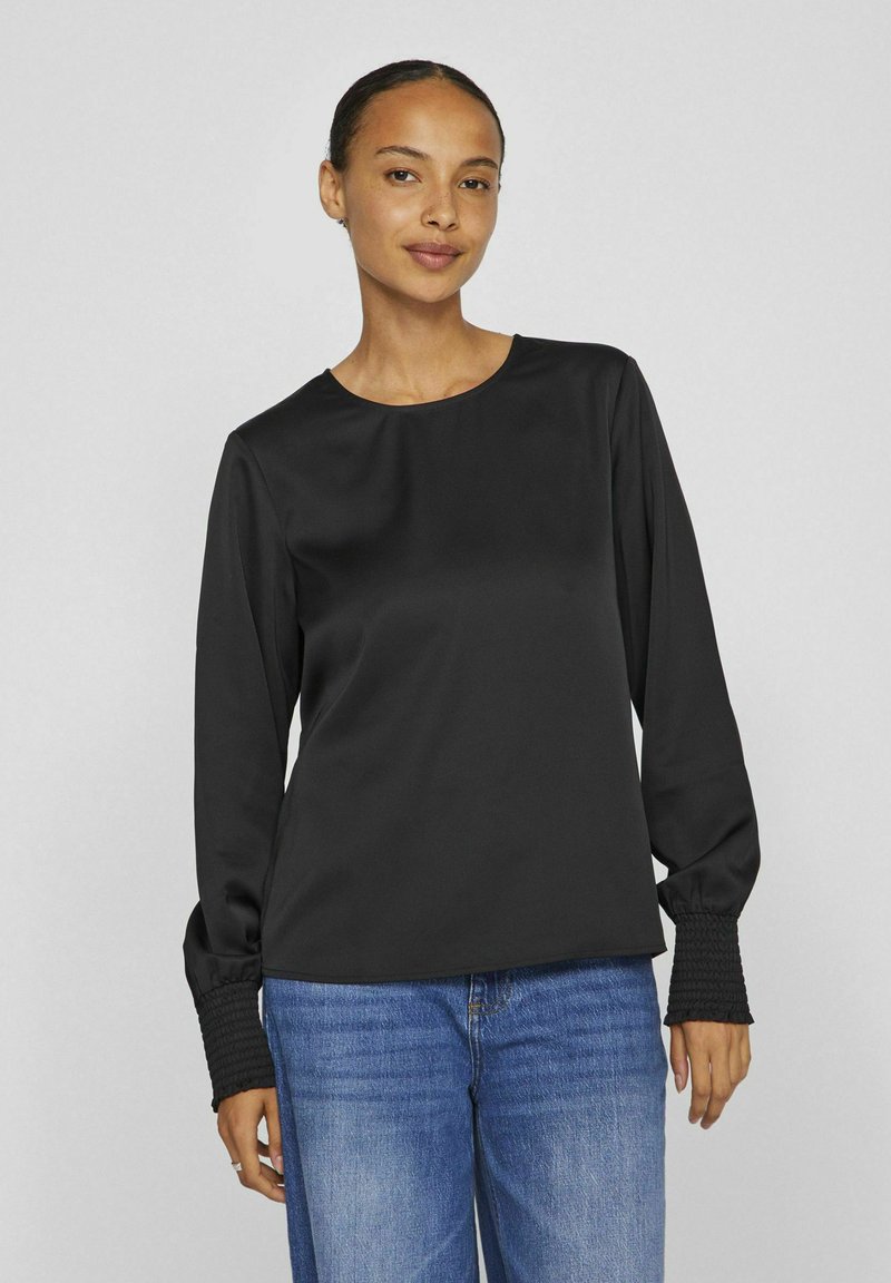 Blusa nera a maniche lunghe con scollo rotondo e polsini testurizzati, realizzata in un tessuto morbido. Abbinata a jeans denim blu chiaro.
