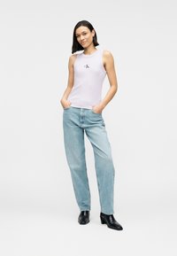 Ανοιχτό μωβ τοπ χωρίς μανίκια με στρογγυλή λαιμόκοψη, διακοσμημένο με ένα διακριτικό κεντητό λογότυπο. Συνδυάζεται με ανοιχτό μπλε παντελόνι φαρδιάς γραμμής και μαύρα μποτάκια με τακούνι.