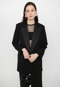 Čierny blejzer so saténovými klopami, nosený cez priesvitný top. Modelka drží malú čiernu kabelku. Výrazné strieborné náušnice dopĺňajú vzhľad.