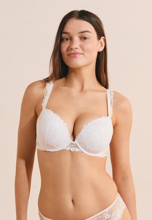 Joven mujer con cabello largo y castaño, vistiendo un sujetador de encaje blanco y ropa interior a juego, de pie contra un fondo beige liso.