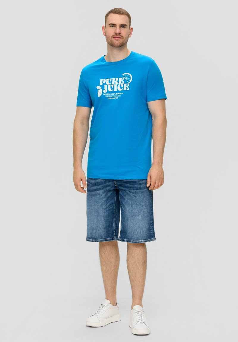 Blauwe katoenen T-shirt met witte opdruk met de tekst "PURE JUICE," gecombineerd met denim shorts en witte sneakers, tegen een lichtgrijze achtergrond.