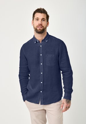 Man with beard and tattoo wearing a navy blue buttoned shirt and light beige pants, standing against a plain light background.