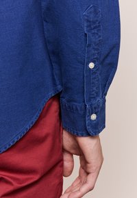 Blå knapp-up skjorta med lång ärm, med en slät yta, rundad fåll och två knappdekorationer på manschetten, kombinerad med röda byxor.