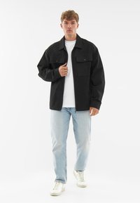 Fekete dzseki elülső zsebekkel, fehér póló fölött viselve, világoskék farmernadrággal és fehér sportcipővel párosítva. Simító anyag és laza fazon.