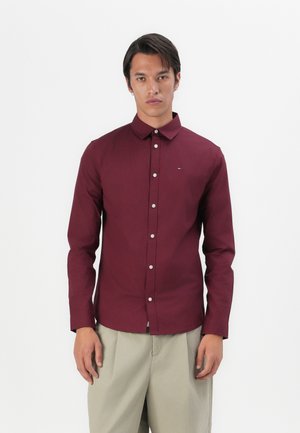 Camisa de manga larga burdeos con cuello abotonado y cierre frontal de botones. Presenta un pequeño detalle de logo en el pecho. Textura suave.