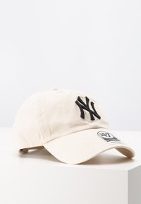 Casquette de baseball en coton beige avec un logo "NY" brodé en noir à l'avant et un patch de marque sur le côté, dotée d'une visière incurvée.