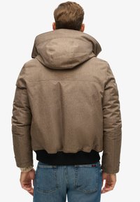 Brun, struktureret pufferjakke med en stor hætte, sort elastisk kant og brandlogo på ryggen. Båret med blå jeans.