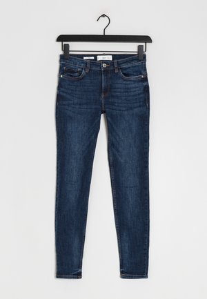 Vaqueros ajustados de color azul oscuro colgados en una percha negra contra un fondo blanco liso.