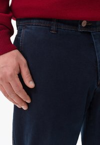Jeans in denim blu scuro con una finitura liscia, caratterizzati da un design frontale semplice e dettagli cuciti. Mano appoggiata sul tessuto.