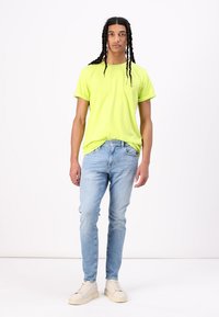 Neon sárga pamut póló, slim-fit világoskék farmernadrág és fehér sportcipő. A modell egyenesen áll egy sima fehér háttér előtt.