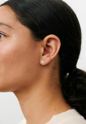 Gros plan de l'oreille gauche d'une femme portant une petite boucle d'oreille clou en forme de fleur argentée, avec des cheveux foncés tirés en queue de cheval.