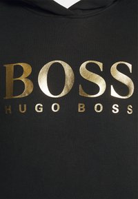 Tissu noir avec un grand texte doré brillant "BOSS" et un texte doré plus petit "HUGO BOSS" en dessous, partie d'un vêtement avec une capuche visible.