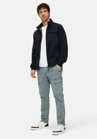 Marineblauwe jas met rits en hoge kraag met gele accenten, grijze cargobroek met zakken, en witte sneakers met marineblauwe accenten.