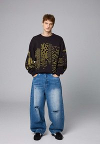 Sudadera negra con patrones geométricos amarillos, combinada con vaqueros de mezclilla azul holgados con áreas desgastadas y tachuelas decorativas. Zapatos negros.