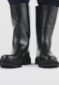 Mustat kumiset sadejalkineet, joissa on sileä pinta, keskipitkät varret, jykevä kuvioitu pohja ja pyöreä kärki.