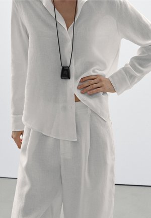 Persoon draagt een losse witte blouse met knopen en bijpassende wijde broekspijpen, met een zwart hangertje aan een koord om de hals.