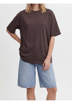 Persona que lleva una camiseta de color marrón oscuro de gran tamaño y pantalones cortos vaqueros azul claro hasta la rodilla, con una mano en la cadera y la otra relajada.