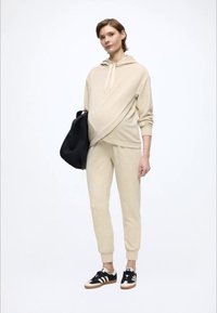 Beige hoodie med korslagd design, matchande beige mjukisbyxor och svarta sneakers med vita detaljer. Bär en svart väska.