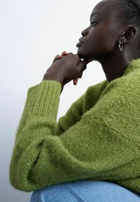 Personne à la peau foncée et aux cheveux courts portant des boucles d'oreilles en argent, un pull en maille vert et un jean bleu, reposant le menton sur les mains croisées de profil.