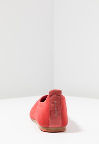 Chaussure plate en daim rouge avec une forme arrondie, une sangle arrière élastique et une semelle lisse. Design minimaliste, sans motifs ni accessoires visibles.