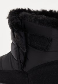 Cizme negre izolate cu o bordură pufoasă din blană neagră în partea de sus. Dispun de un amestec de materiale netede și texturate cu o teamă cu arici.