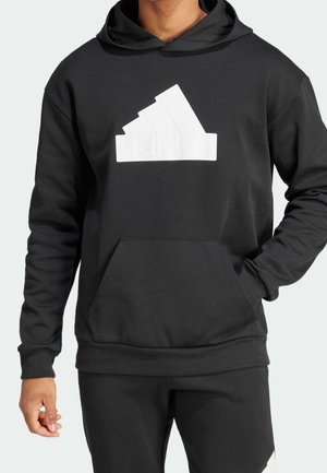 Mężczyzna w czarnej bluzie z kapturem z białym logo Adidas, z kieszenią z przodu i dopasowanymi czarnymi spodnie, stoi z jedną ręką w kieszeni.