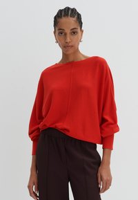 Pull rouge surdimensionné avec un col bateau, des manches longues et une texture douce. Associé à un pantalon fuselé marron foncé.