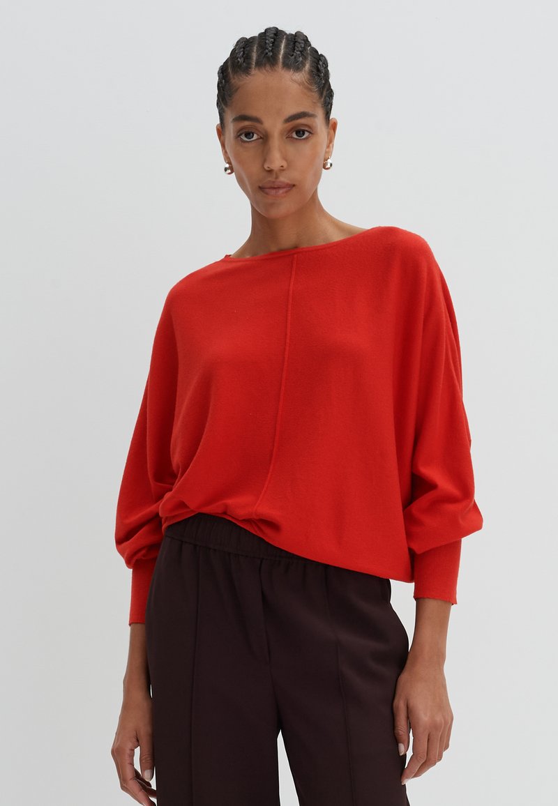Pull rouge surdimensionné avec un col bateau, des manches longues et une texture douce. Associé à un pantalon fuselé marron foncé.