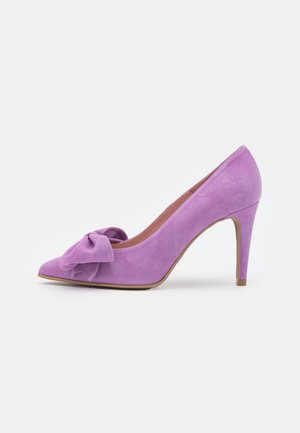 Zapato de tacón alto de ante morado con punta afilada y detalle de lazo de tela en la parte delantera. Presenta un tacón stiletto delgado.