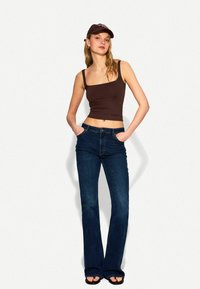 Smeđi krojeni crop top s tankim trakama, uparen s tamnoplavim širokim trapericama i crnim sandalama. Smeđa kapa upotpunjuje odjevnu kombinaciju.