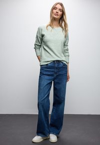 Młoda kobieta stoi ubrana w jasnoszary sweter o fakturze, szerokie niebieskie dżinsy i białe trampki, na tle gładkiej białej ściany.