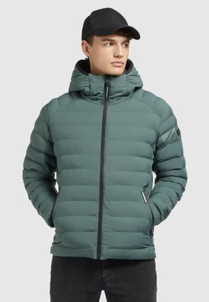 Gepolsterte grüne Jacke mit Kapuze, ausgestattet mit horizontalen Nähten, einem Frontreißverschluss und zwei Seitentaschen. Getragen mit einer schwarzen Mütze.