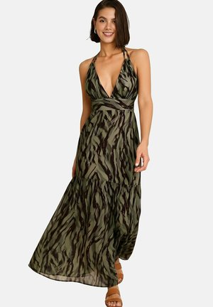 Femme souriante portant une robe maxi verte à imprimé tigre sans manches, avec un col en V, associée à des sandales plates couleur beige sur fond uni.