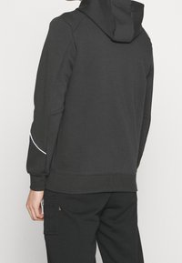Sudadera negra con un patrón texturizado en los hombros, bolsillo tipo canguro y un acento blanco en la manga. El material parece suave y elástico.