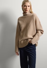 Maglione beige a coste con collo alto, caratterizzato da linee di texture diagonali; abbinato a jeans a gamba larga blu scuro. Tessuto liscio, vestibilità comoda.