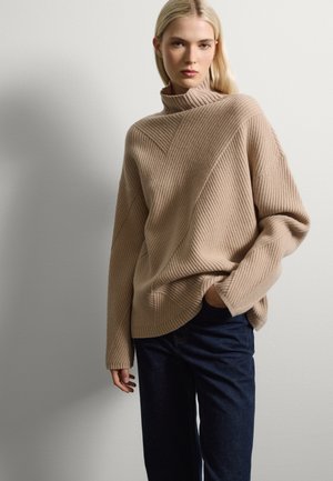 Beige gerippter Pullover mit hohem Kragen, der diagonale Strukturlinien aufweist; kombiniert mit dunkelblauen weiten Jeans. Glattes Material, lässiger Schnitt.