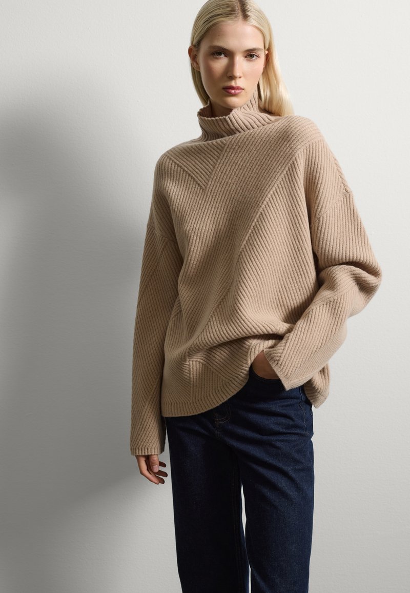 Maglione beige a coste con collo alto, caratterizzato da linee di texture diagonali; abbinato a jeans a gamba larga blu scuro. Tessuto liscio, vestibilità comoda.