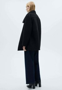 Black wool coat with a structured collar, oversized silhouette, and folded lapels. Paired with wide-leg dark denim jeans and heeled boots.